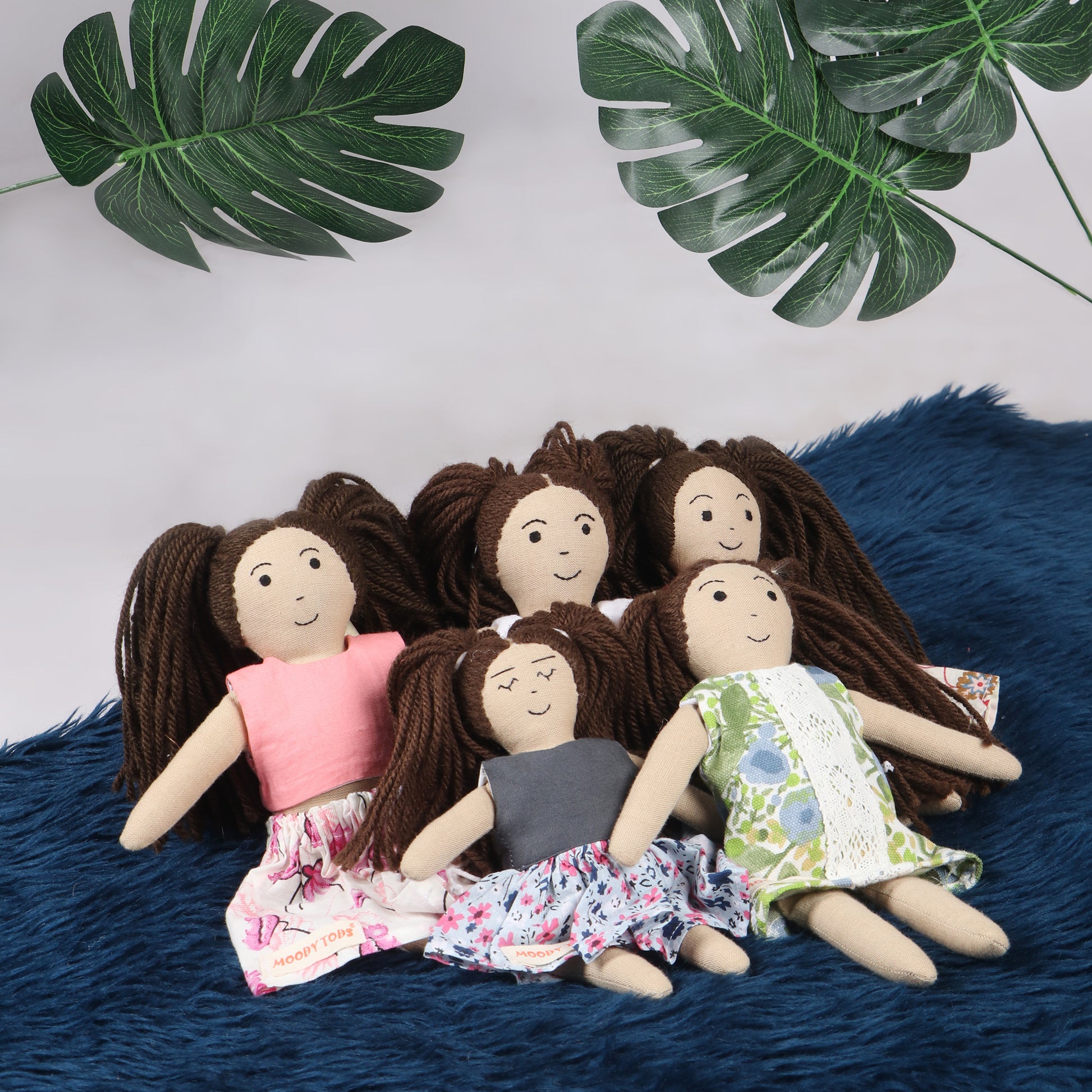 Side view of 5 cute handmade girl rag dolls in sitting position with green leaves in the background made from child-safe, soft fabric