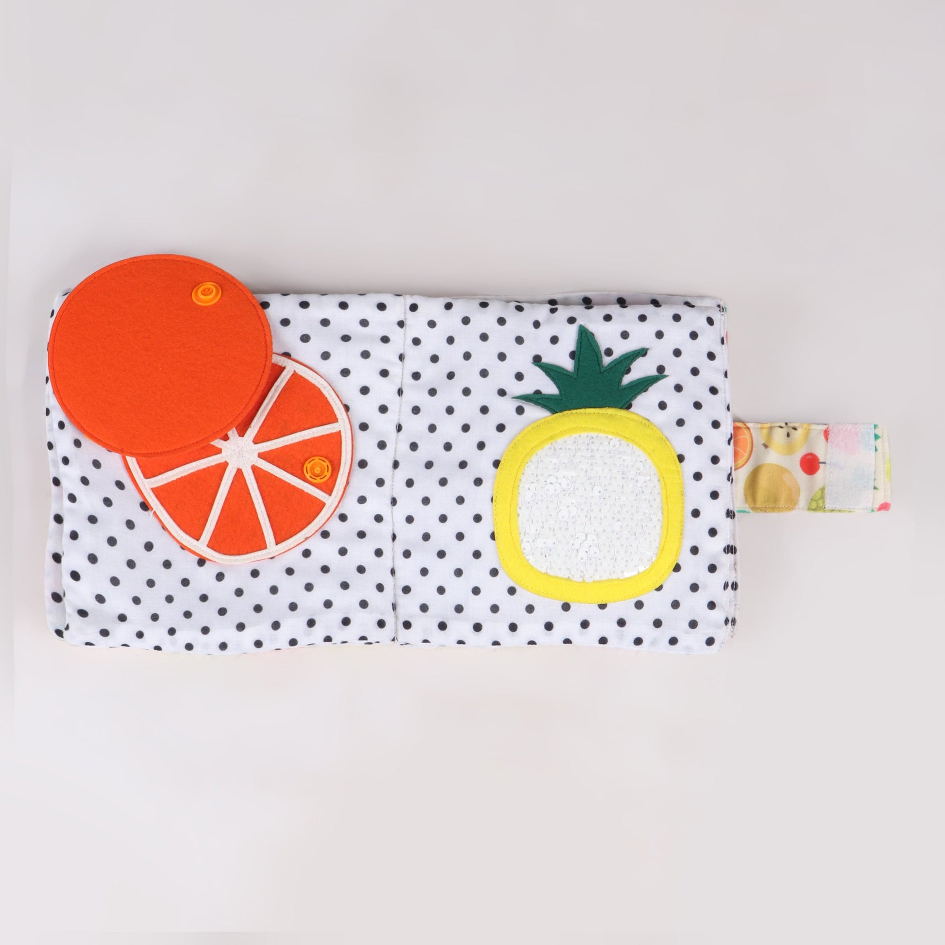Interactive fabric fruit book featuring a push button on an orange to reveal its inside and sequins on a pineapple to depict its textured surface, designed for sensory exploration and fine motor skill development