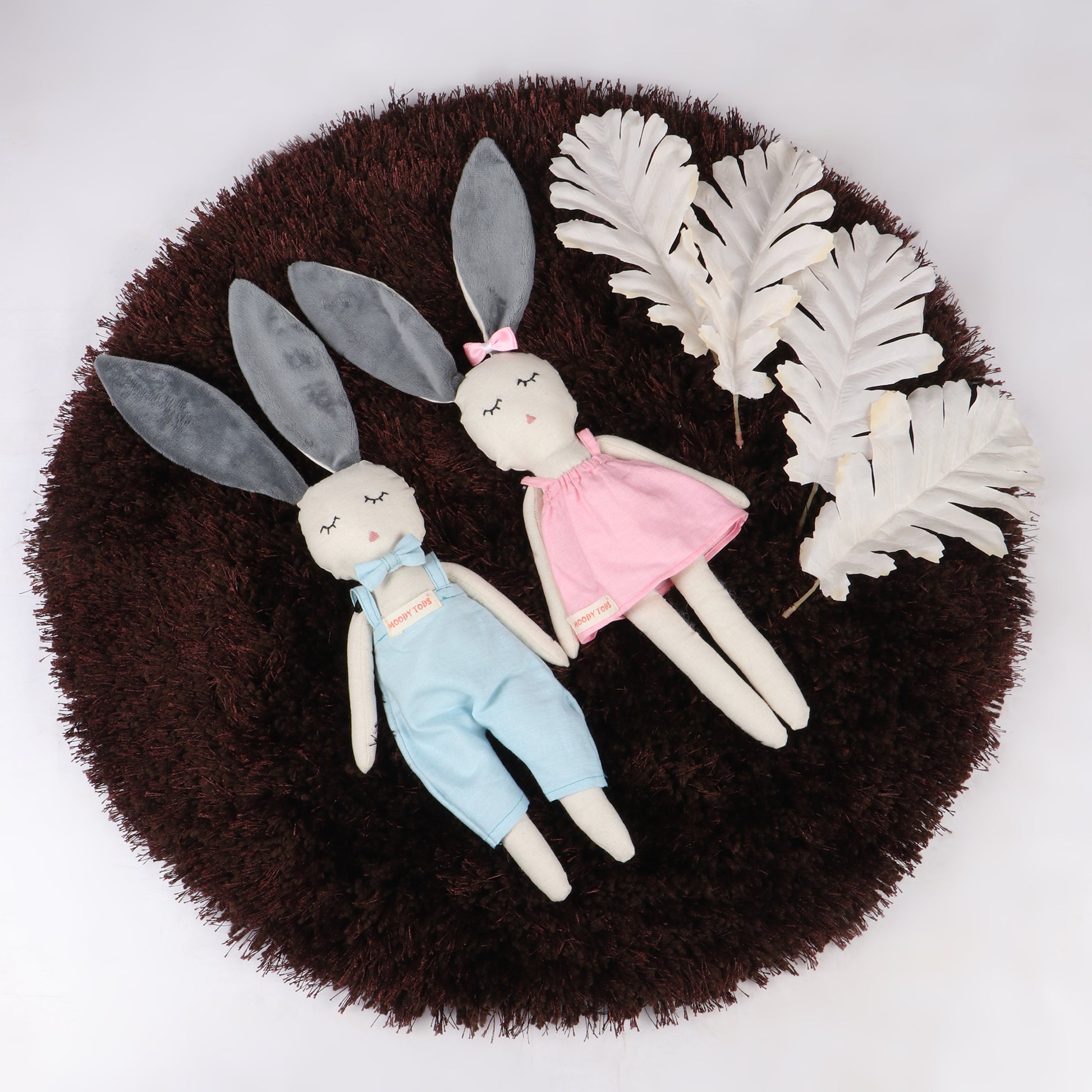 Pair of an adorable 12 inch plush bunny toys with blue and pink pastel colored outfit, perfect for cuddles and sensory play lying flat on brown colored rug
